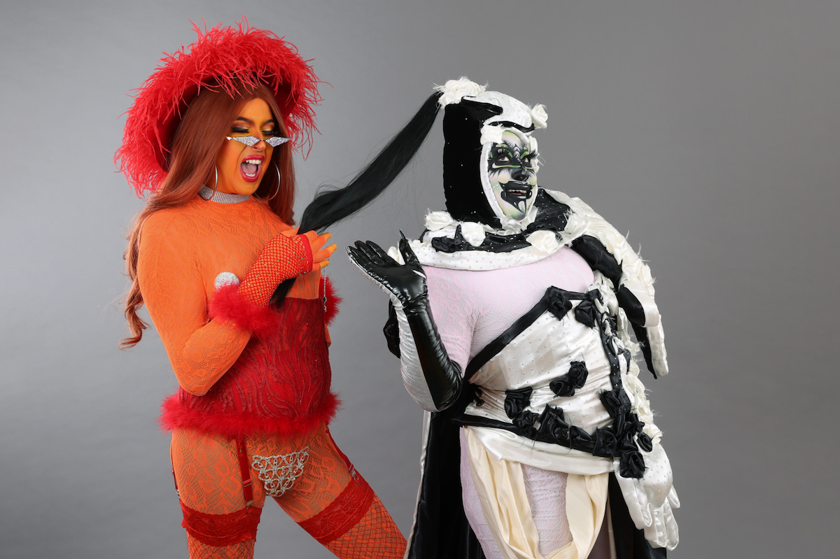 A drag artist in an orange and red feathery outfit poses with another drag artist wearing black-and-white abstract designs. 