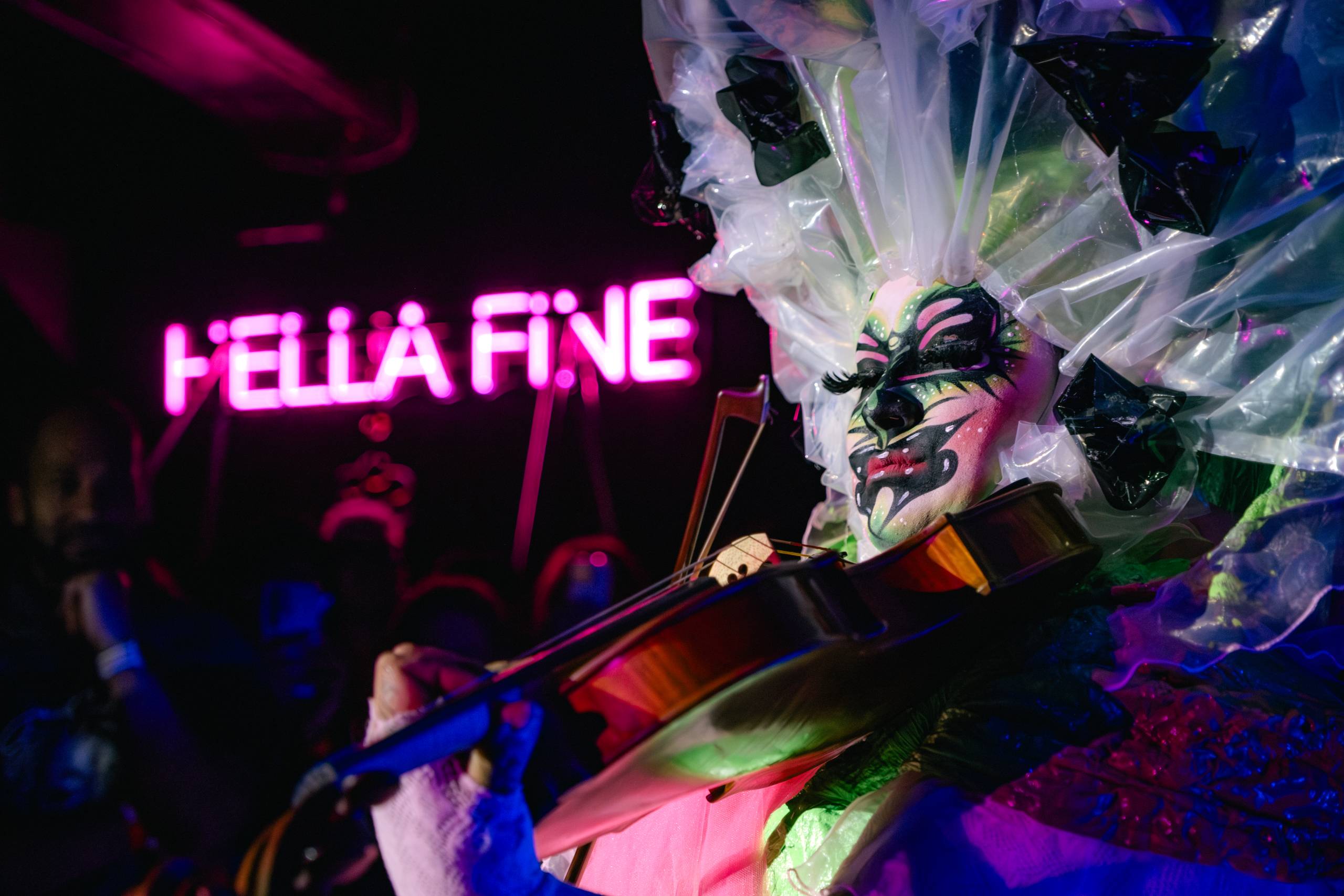 A drag artist plays a viola inside a bar with neon text behind them that says "Hella Fine."