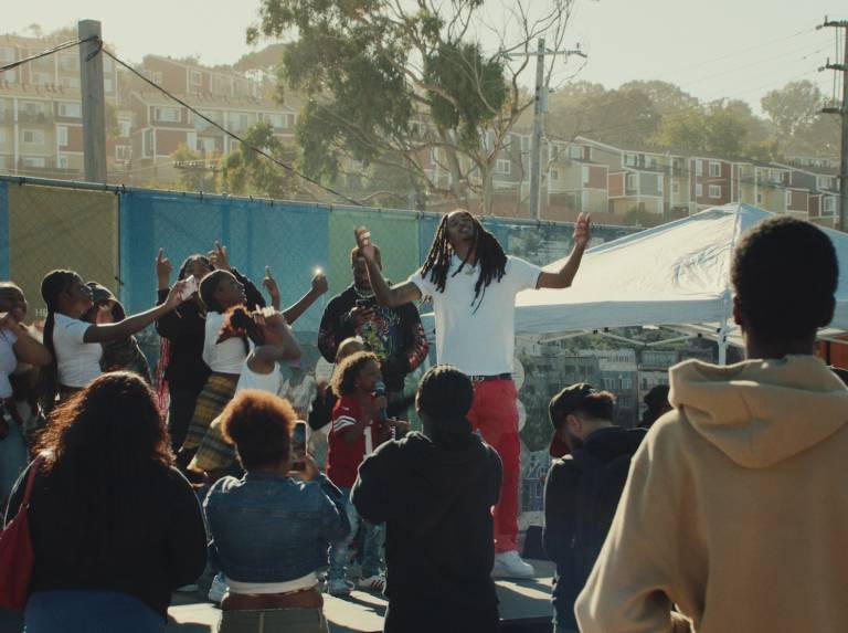 A man on an outdoors stage in a white shirt has his hands lifted in the air as he stands in a circle of kids with one of the children singing into a microphone.