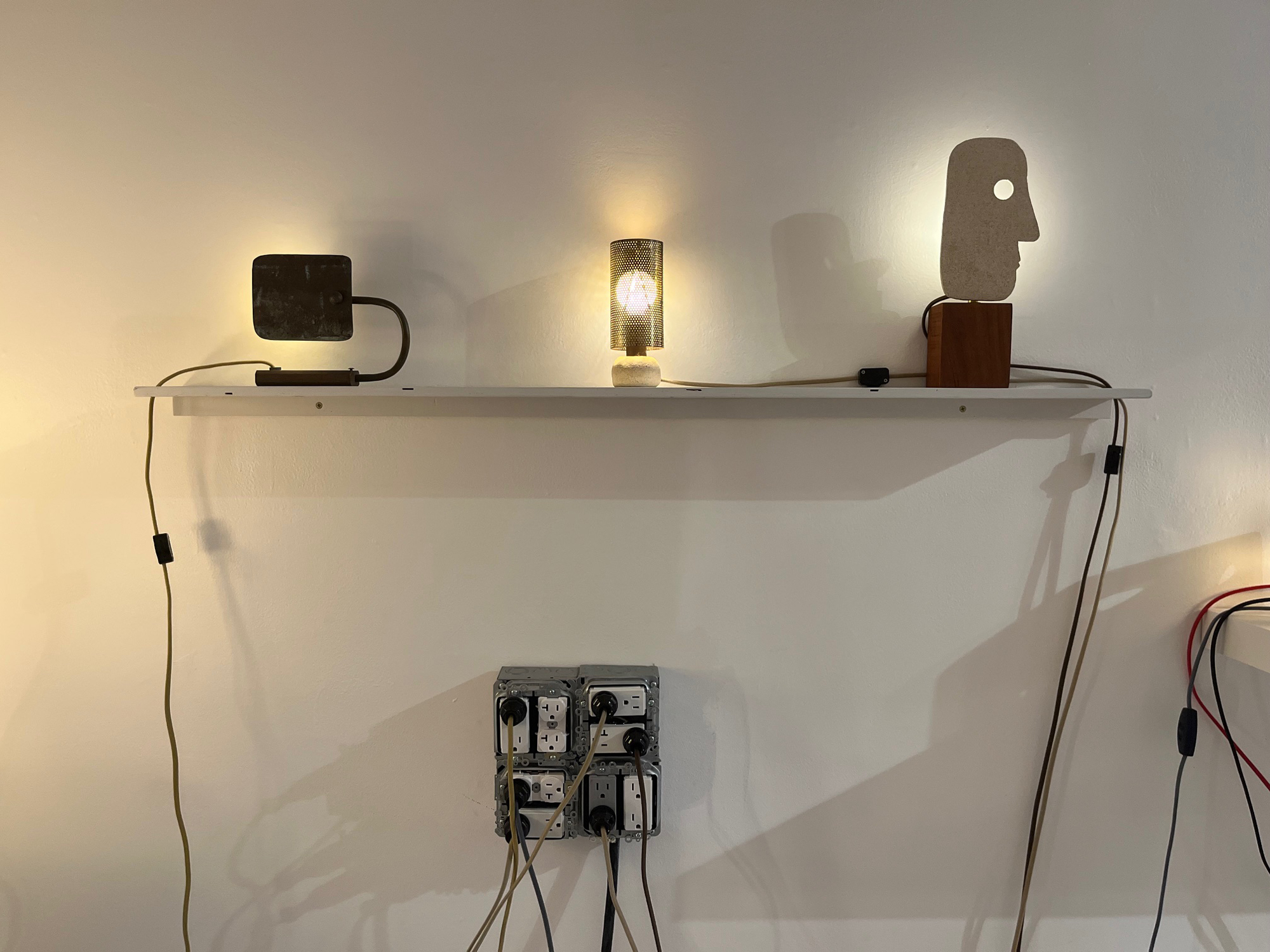 three artistic lamps on shelf above a beefed-up outlet with plugs in it