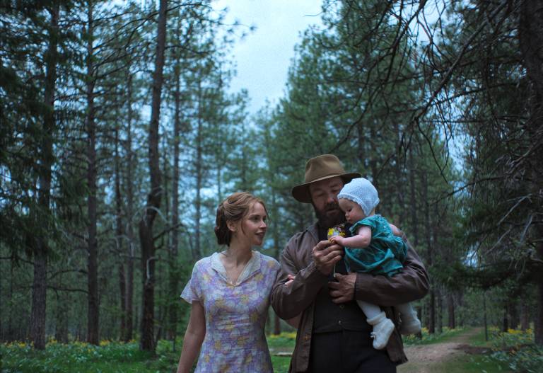 A rural-dressed young couple walk through the woods with a newborn baby