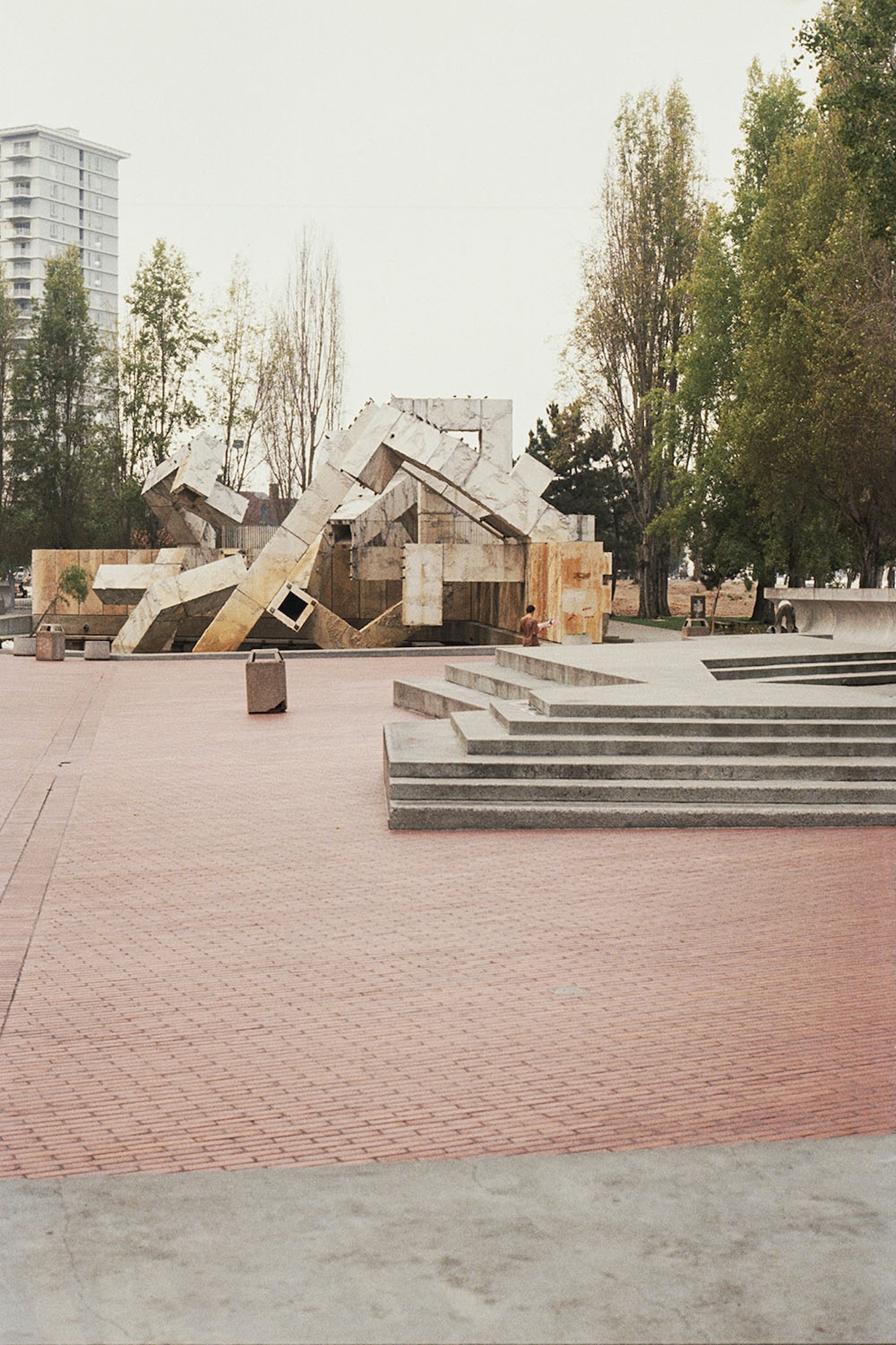 A brutalist concrete foundtain constructed from snaking square pipes is viewed on an overcast day. Two tall green trees stand next to the structure.