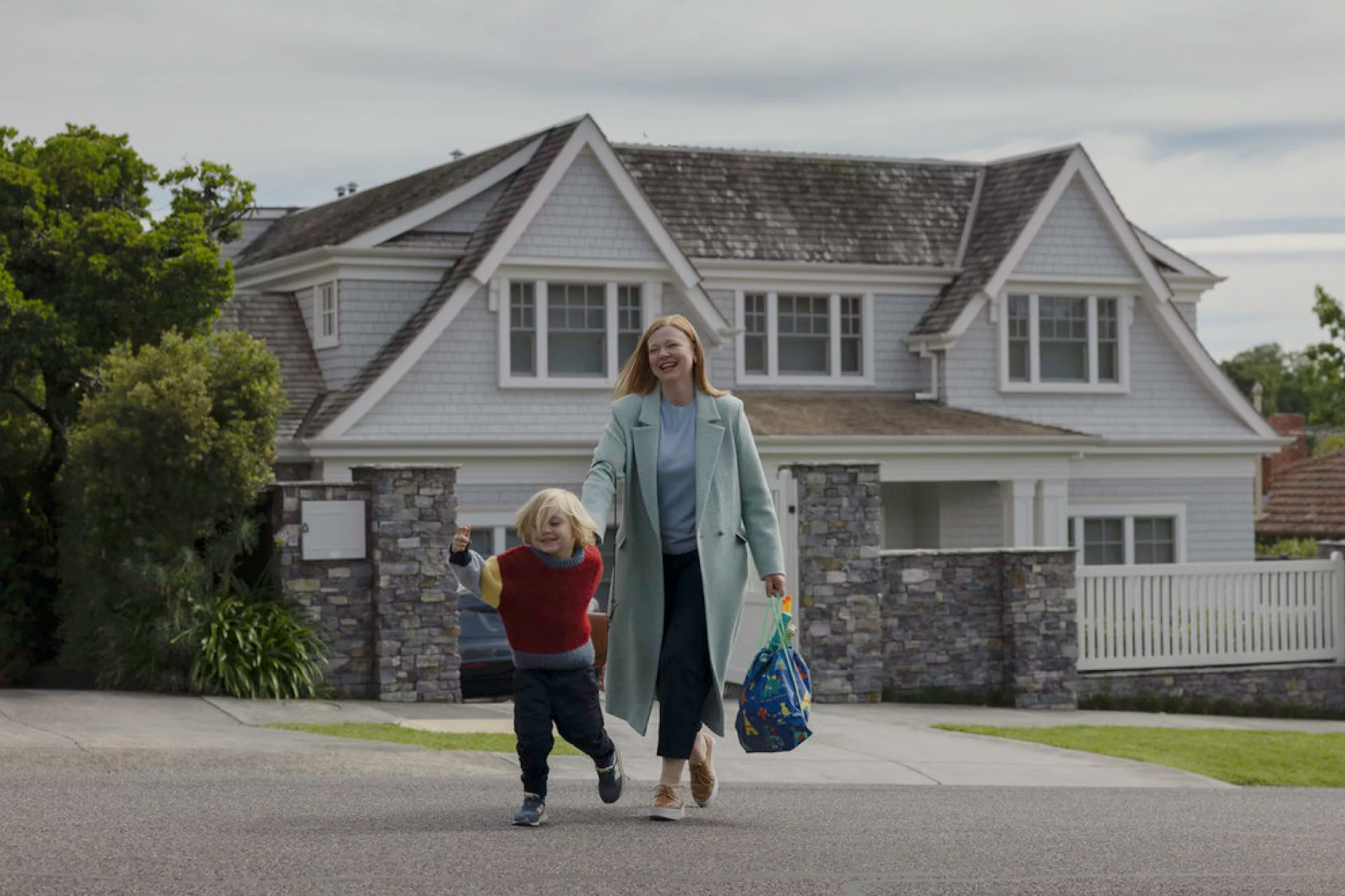 A white middle-aged woman dressed in smart casual clothes walks a small blond child across the street from a mansion. She is smiling and carrying a children's bag.