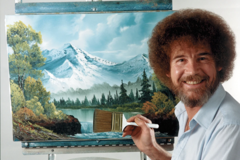 A smiling white man with bushy brown hair and beard, wearing a pale blue shirt, stands next to a painting of a mountain top and countryside, paintbrush in hand.