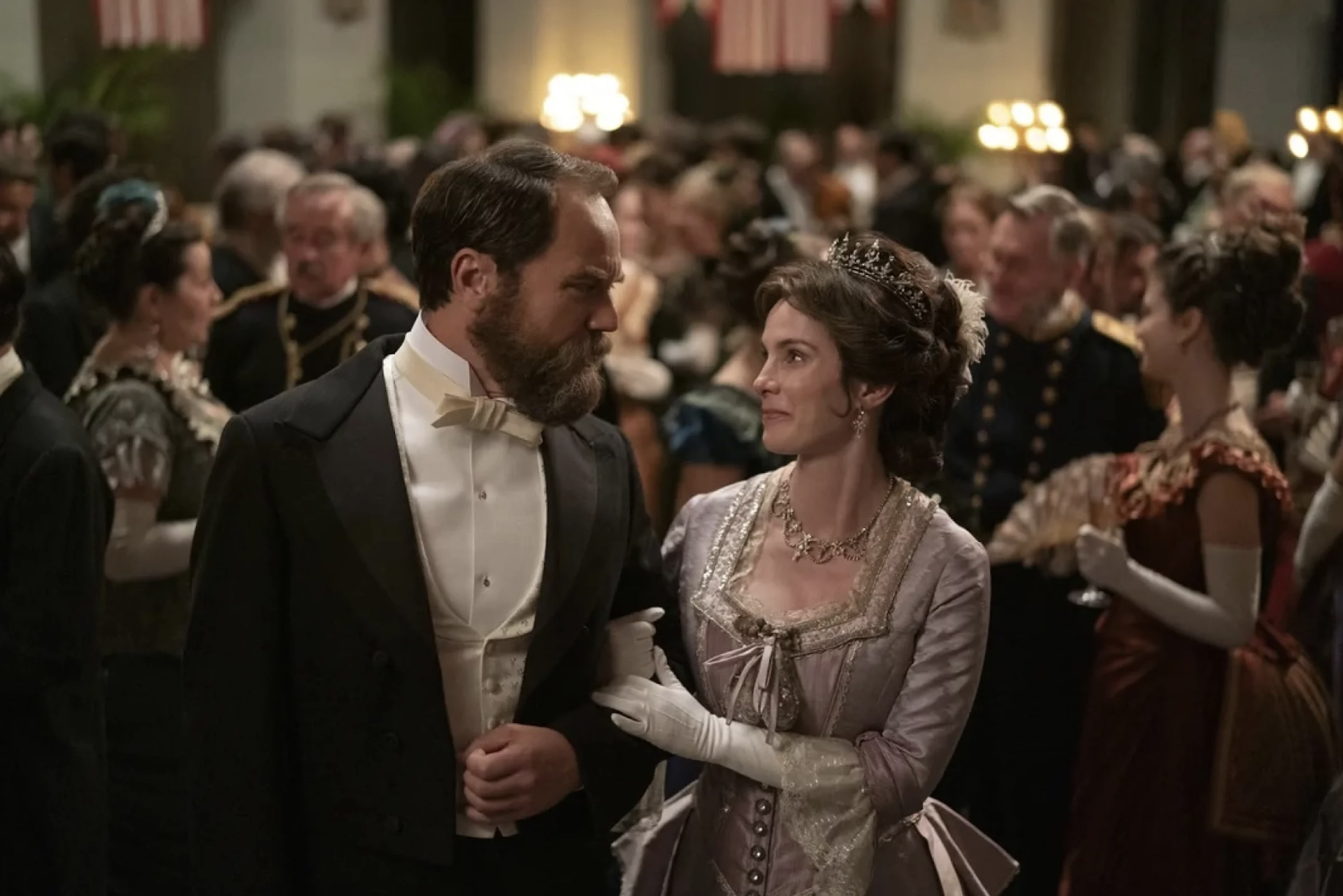 A white man and woman dressed in period costume stand, arms linked, in the middle of a packed ballroom, looking at each other affectionately.
