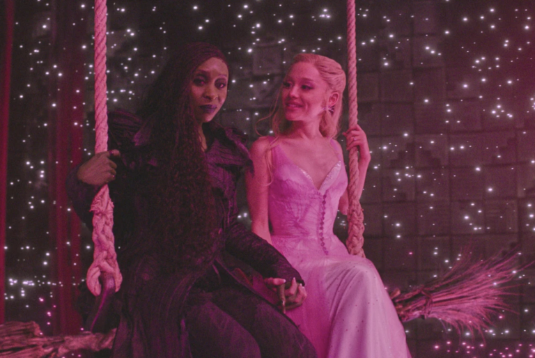 Two women — one dainty, white, wearing a strapless gown and smiling; the other worried-looking, wearing heavy black cloaks, her face and hands green — sit side by side on a large swing.