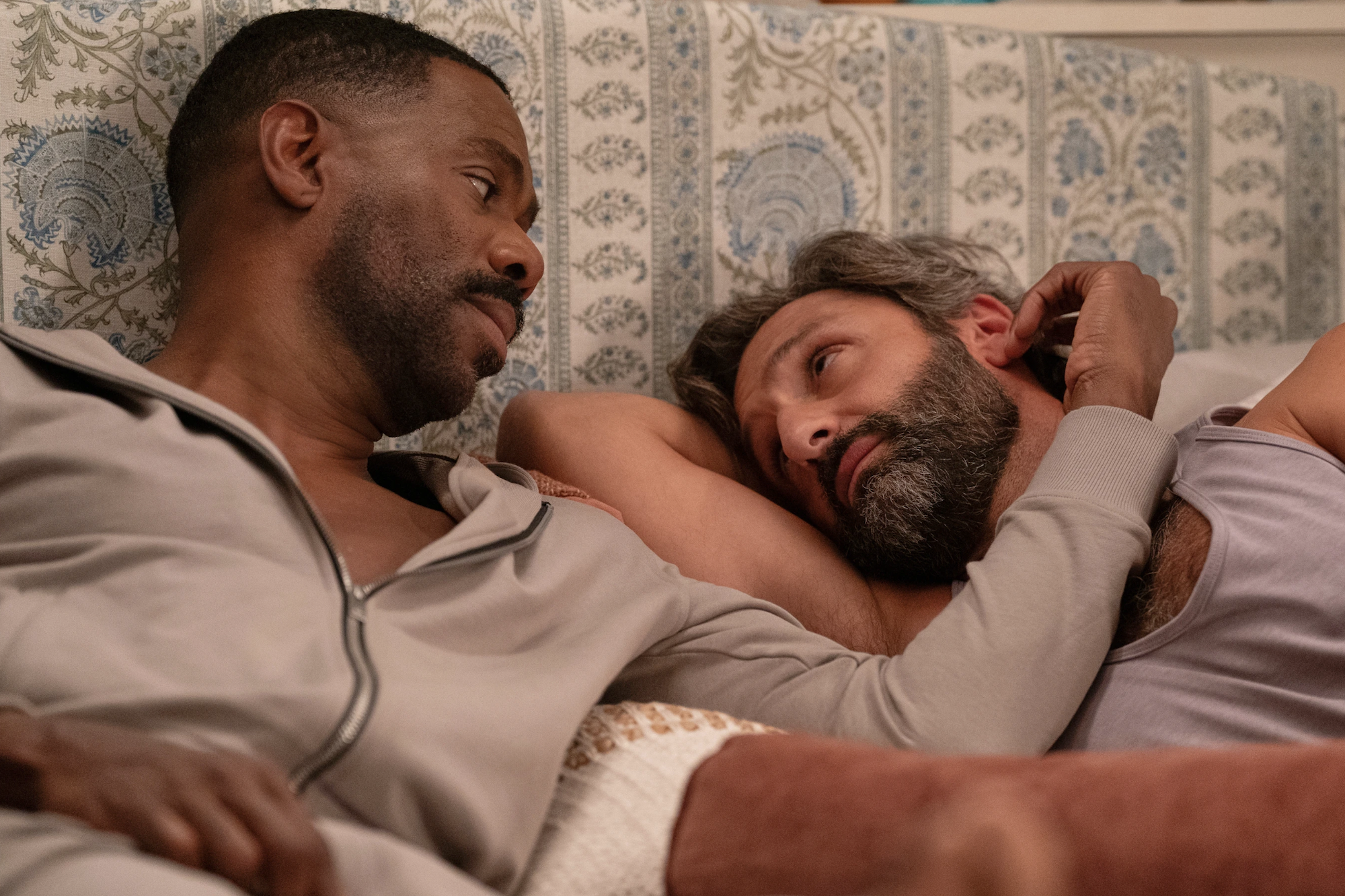 A Black man and a dark-skinned white man with a beard recline in bed together peacefully. 