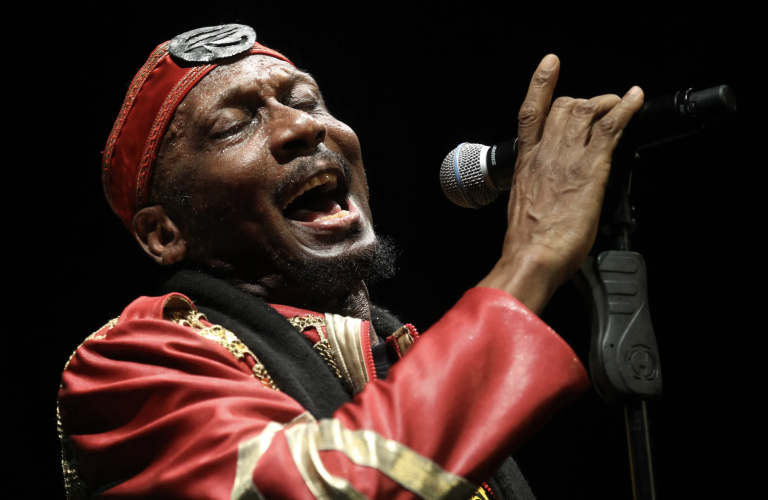 An older Black man singing passionately, eye closed, into a microphone on stage. Hr is wearing a red leather jacket and matching headband.