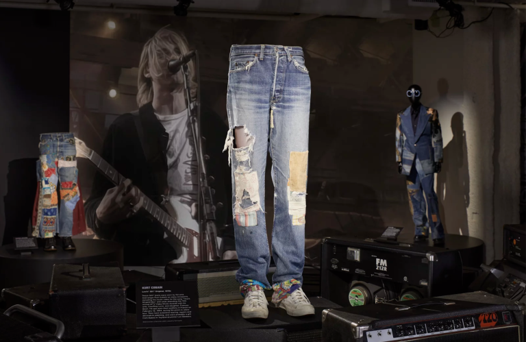 A pair of ripped and patched-up blue jeans stand on mannequin legs in the center of a display, in front of a photo of Kurt Cobain from Nirvana, performing on stage.