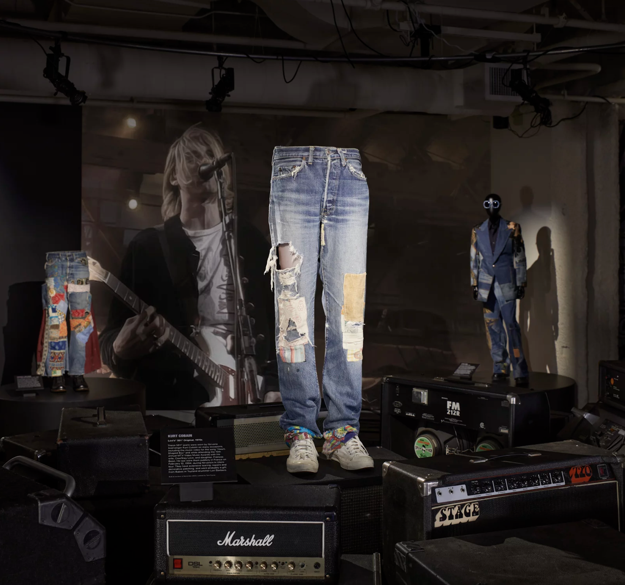 A pair of ripped and patched-up blue jeans stand on mannequin legs in the center of a display, in front of a photo of Kurt Cobain from Nirvana, performing on stage.