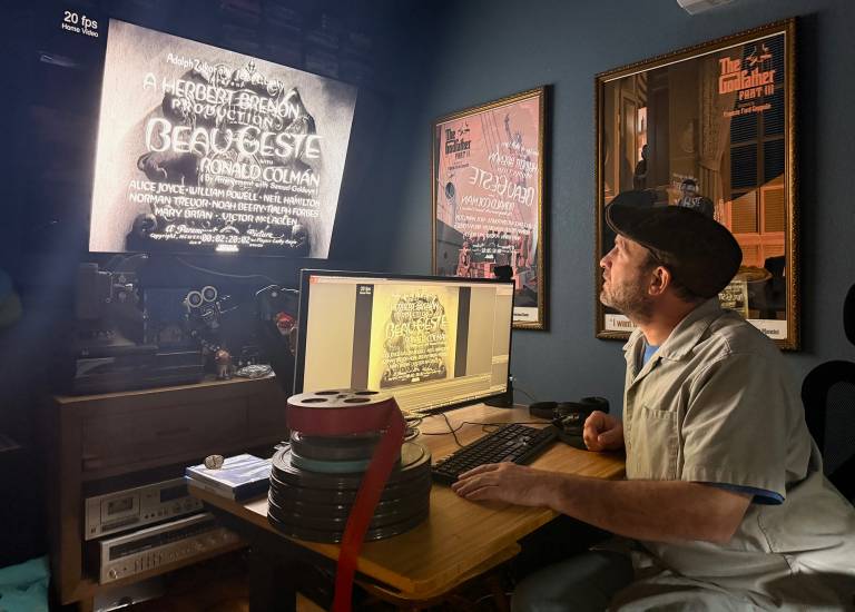 A man in a grey shirt and dark cap sits at a desk with a computer screen, showing a black and white film title, looking up at a second screen higher on the wall, surrounded by movie posters and vintage film and stereo equipment..