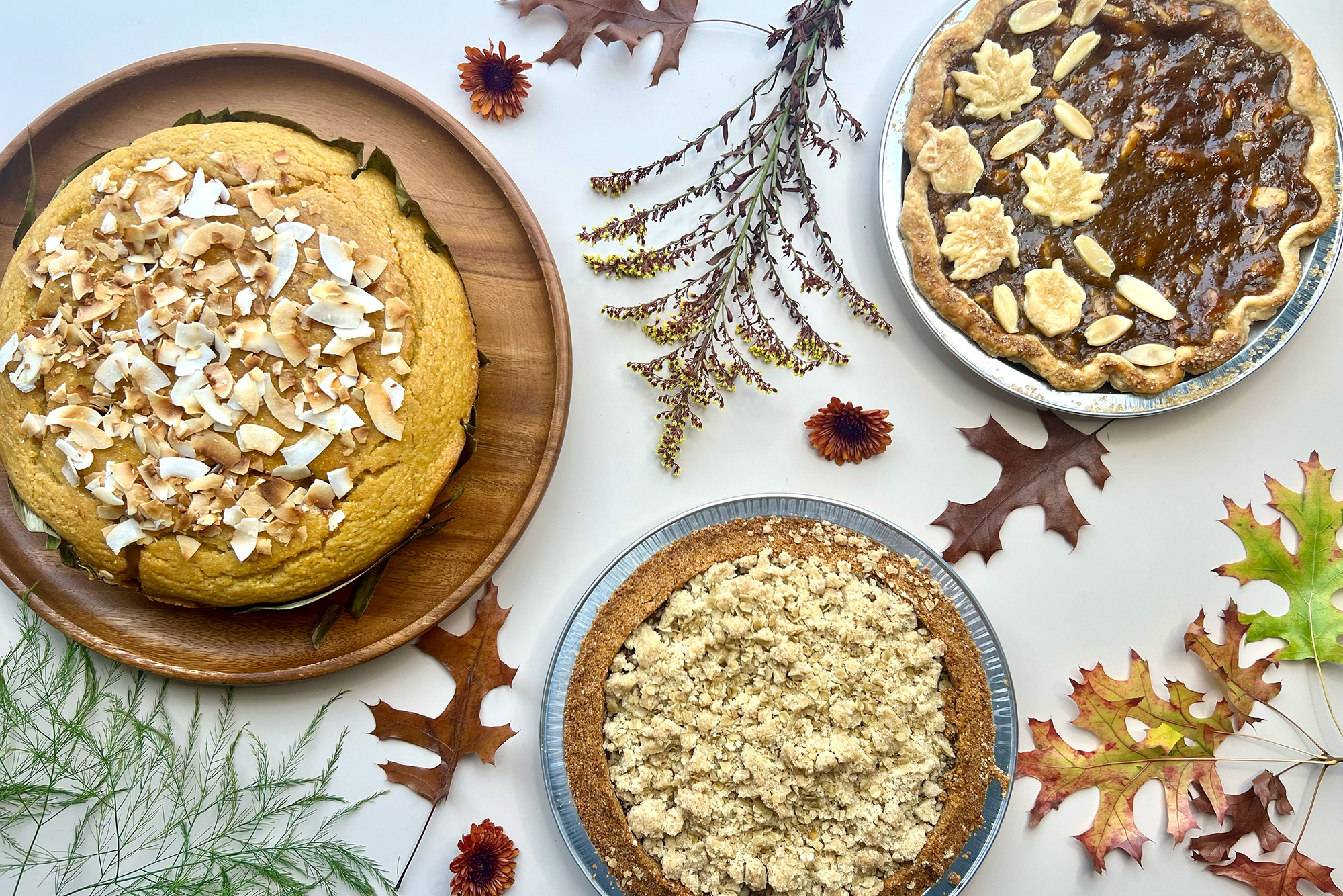 A spread of desserts on a festival holiday table decorated with autumn foliage. There are two pies and a cornbread-like cake topped with toasted coconut flakes.