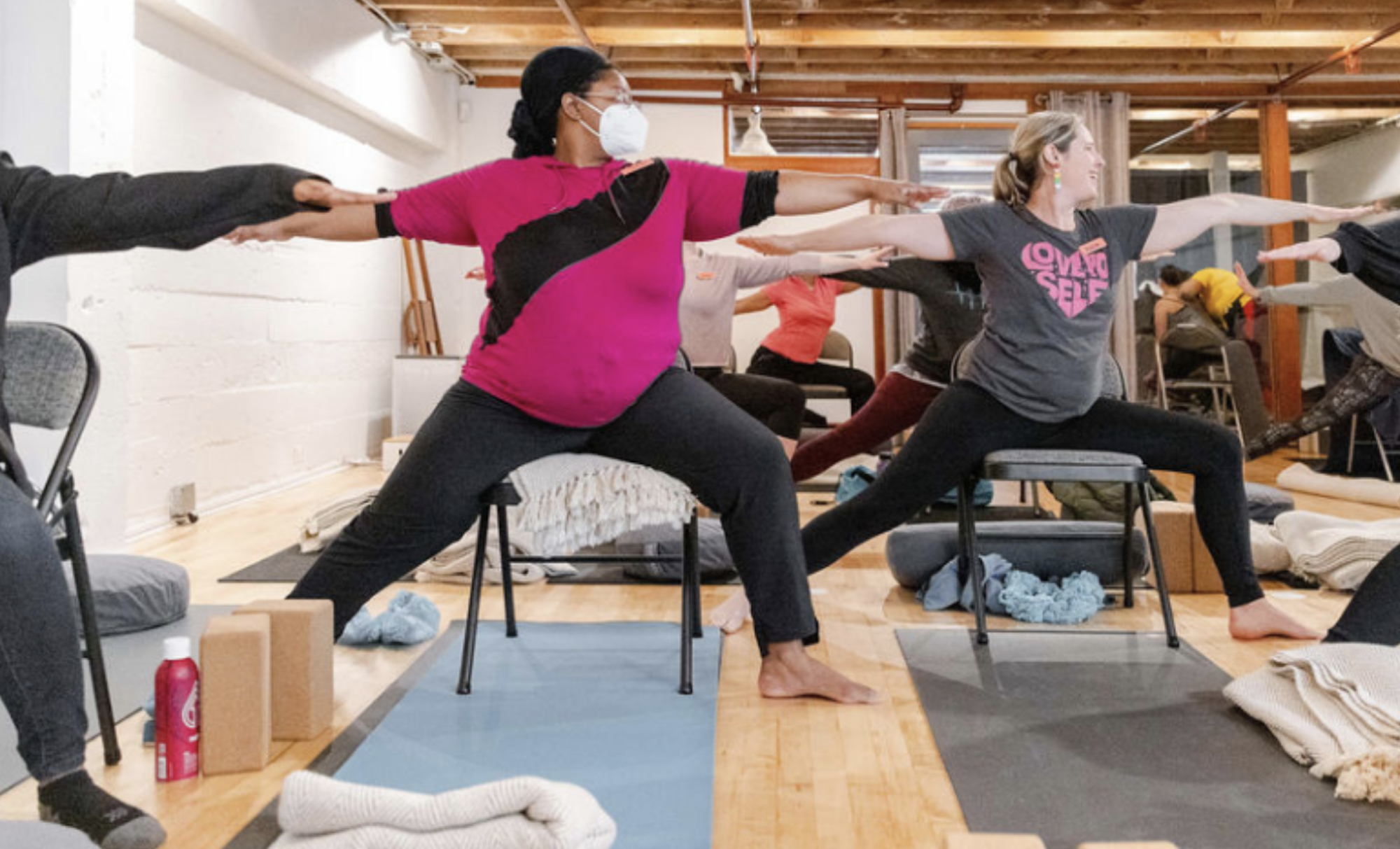 Three women in relaxed clothes perform sideways yoga stretches, aided by chairs.
