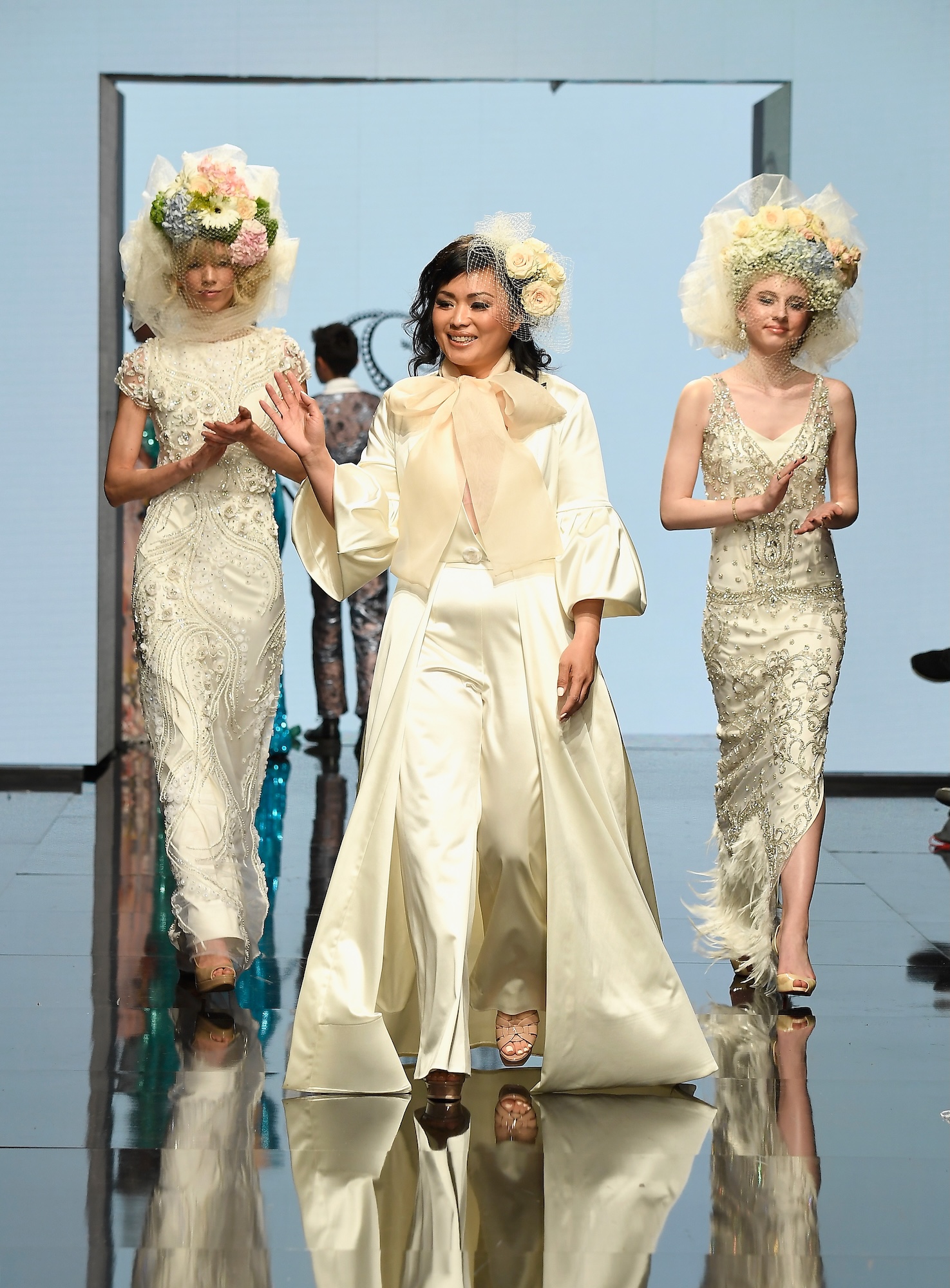A fashion designer in a white, silky outfit walks the runway next to two models in white silk dresses with elaborate headpieces. 
