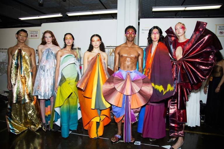 Backstage at a fashion show, models line up wearing colorful geometric gowns.