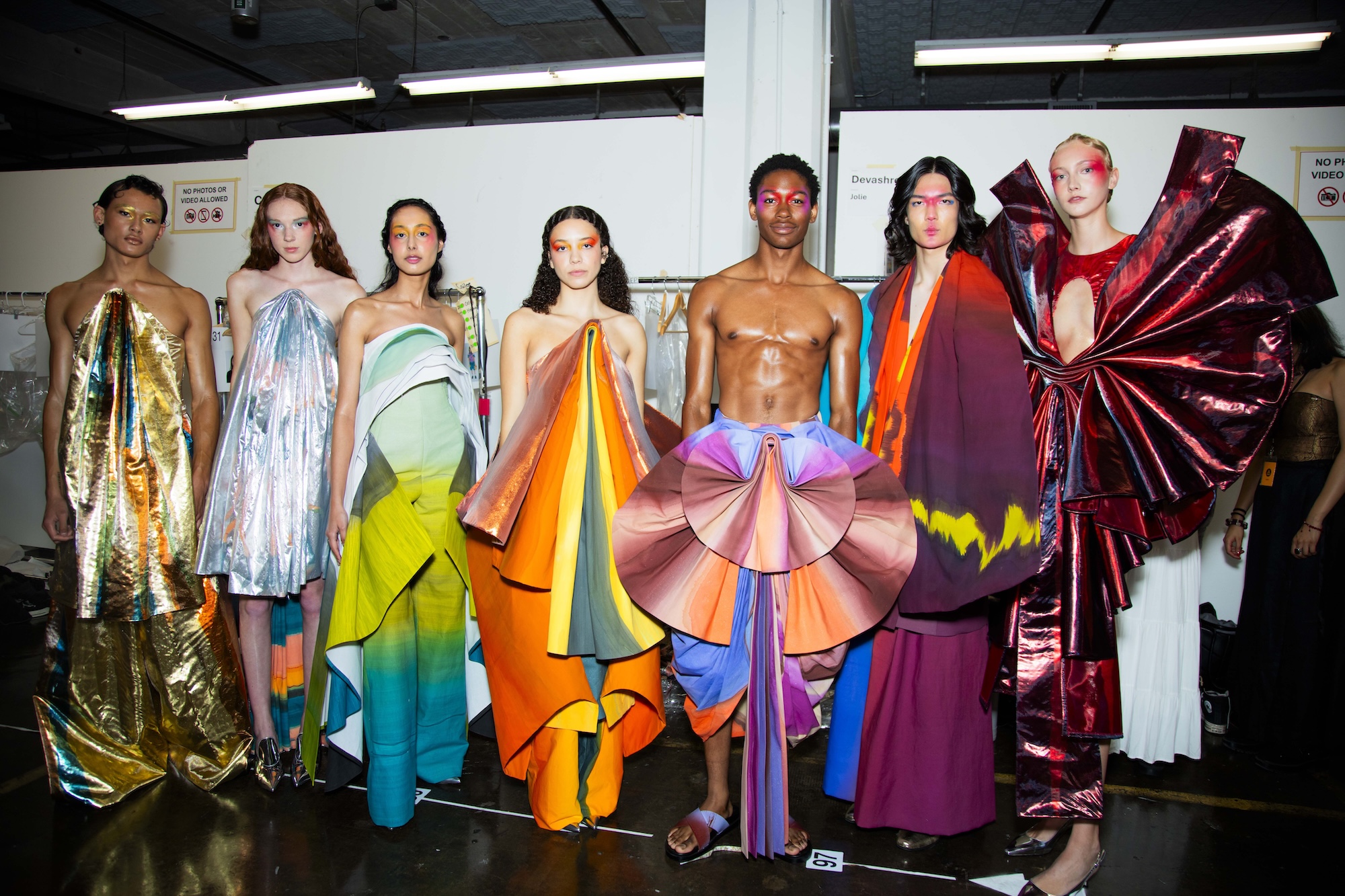 Backstage at a fashion show, models line up wearing colorful geometric gowns.