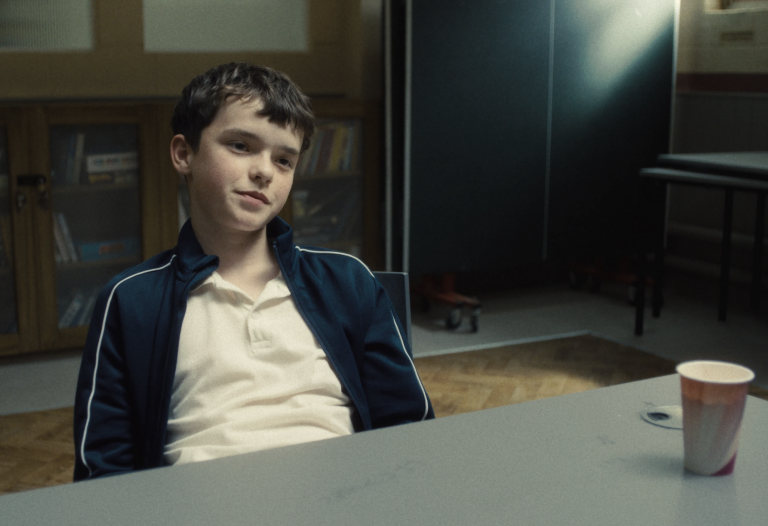 A boy of around 13 sits in a chair at a table with a disposable drink cup on it. He is wearing a white polo shirt and casual jacket. He looks unimpressed.