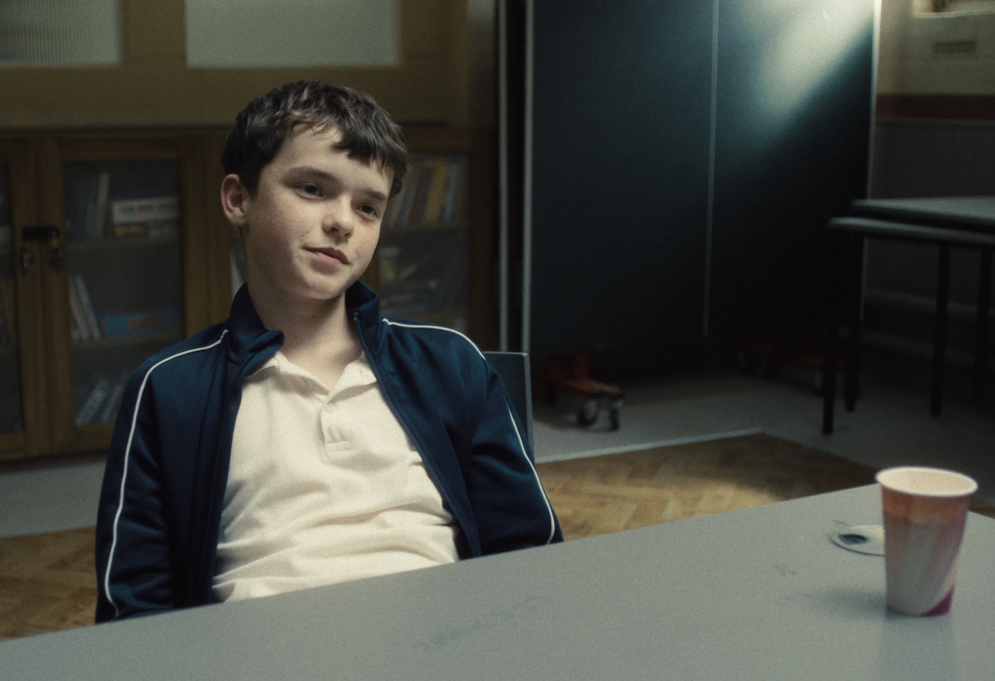 A boy of around 13 sits in a chair at a table with a disposable drink cup on it. He is wearing a white polo shirt and casual jacket. He looks unimpressed.