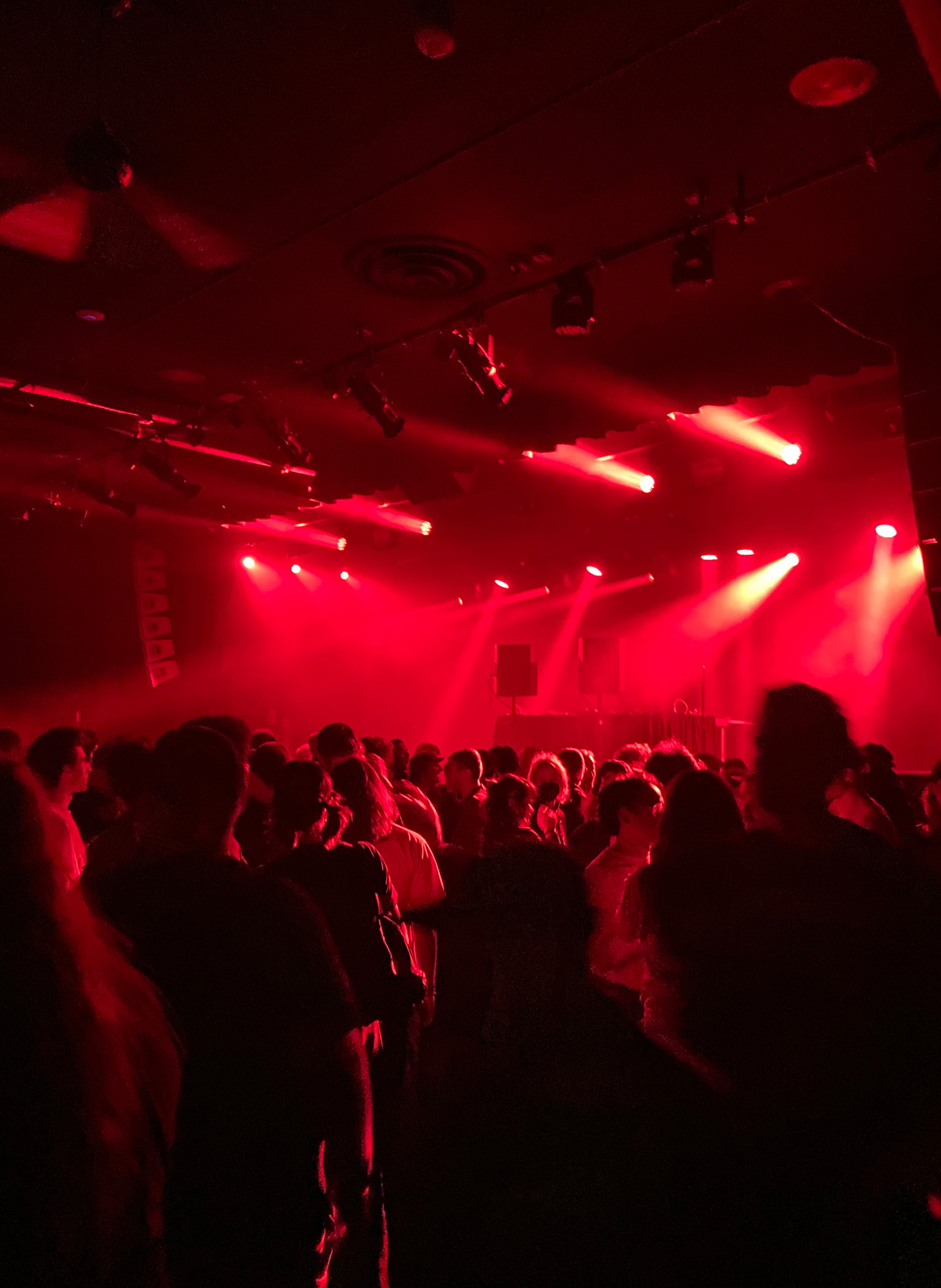 Red lights shine on a crowd seen in backlit silhouettes, with a stage in the distance