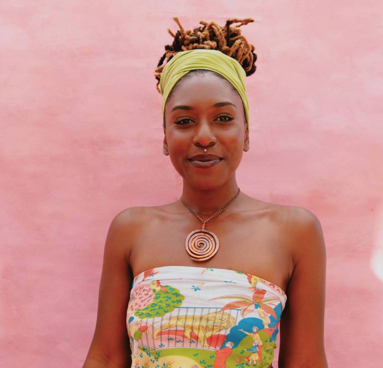 An African-American woman poses for a photo in front of a pink background, she's wearing a white and yellow top with a khaki skirt.