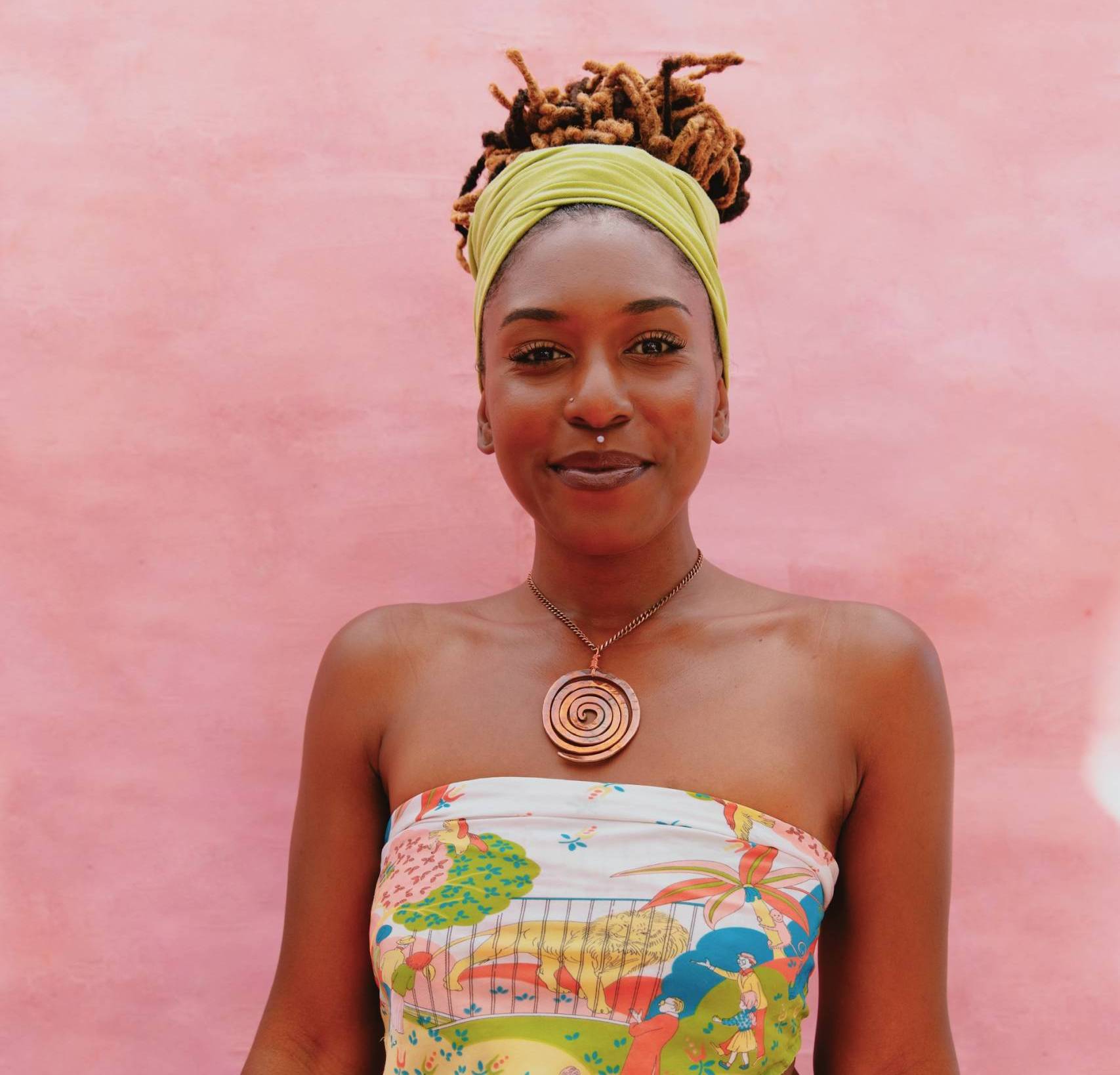 An African-American woman poses for a photo in front of a pink background, she's wearing a white and yellow top with a khaki skirt.
