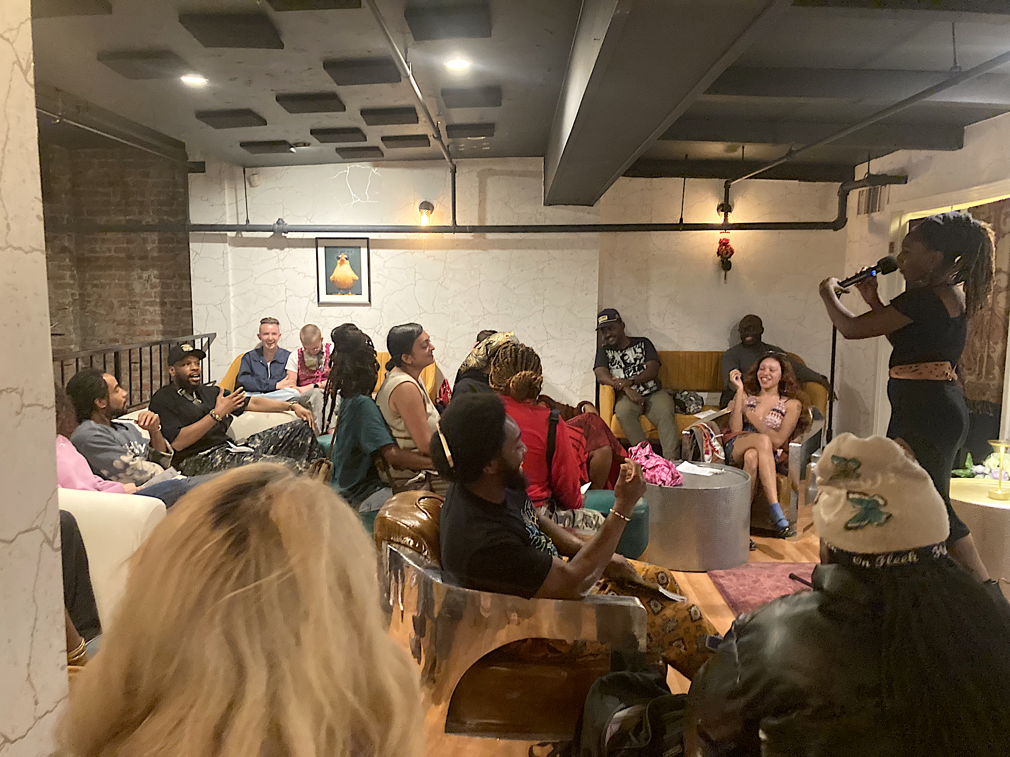 An African-American woman with shoulder-length locs and all black attire stands on a stage in an intimate room as she hosts and open mic event.