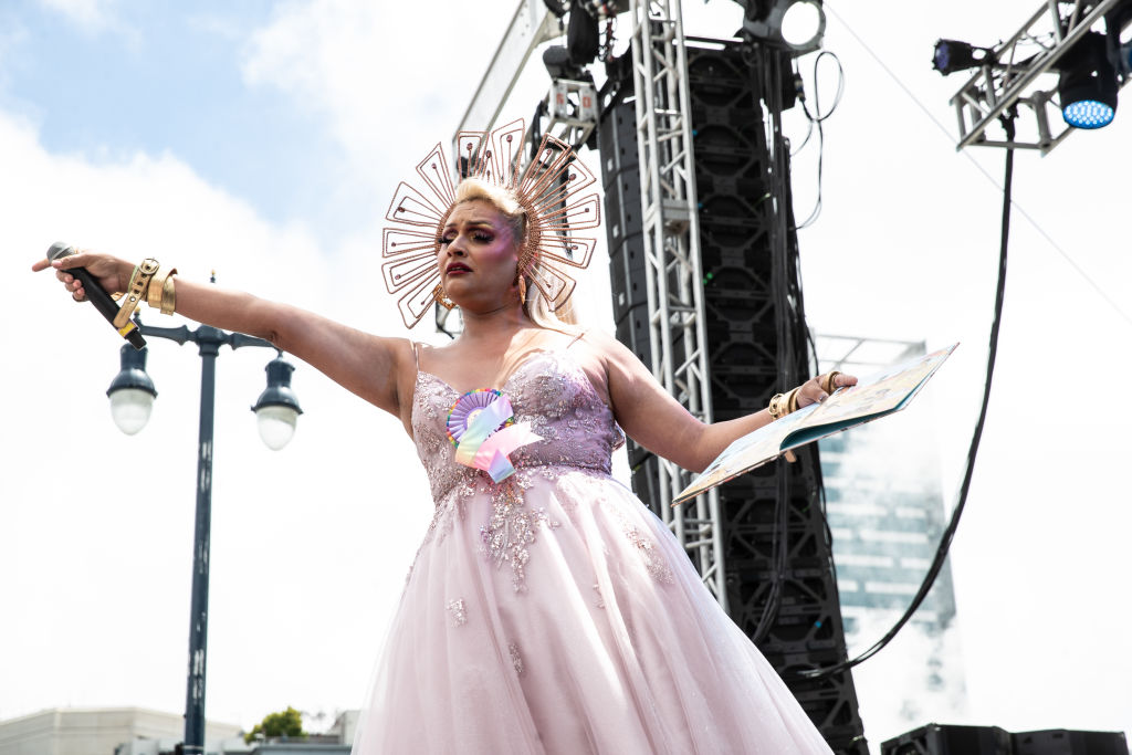 A drag queen in a light-pink gown holds a children's book on stage.