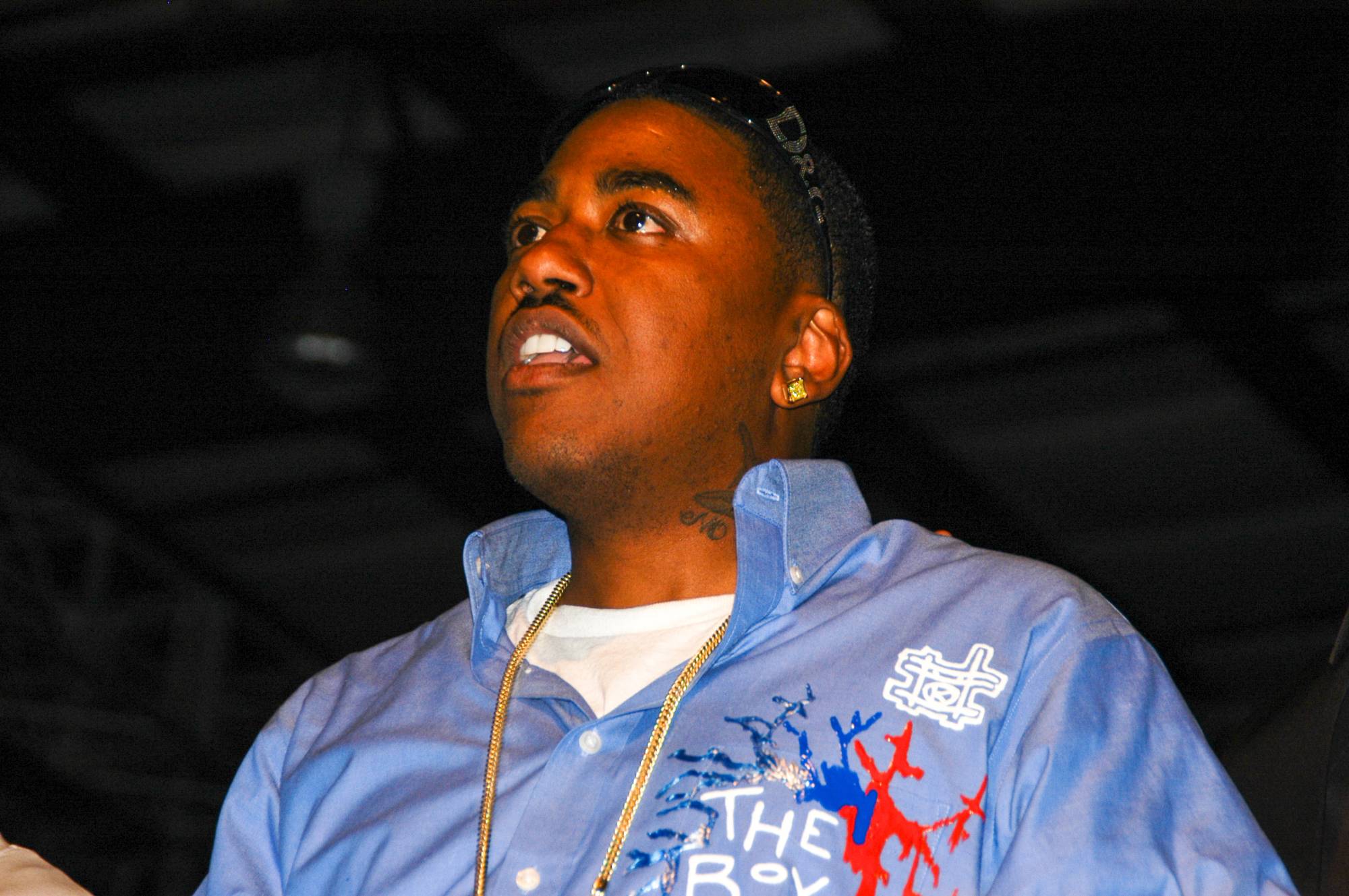 A twentysomething Black man in a light blue collared shirt, gold chain and sunglasses on top of his head looks upward and to the left