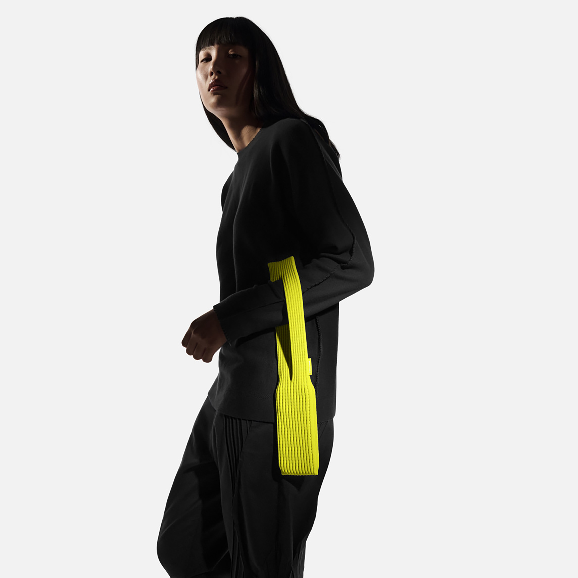 An Asian woman with long black hair and a loose-fitting charcoal top and pants holds a neon yellow thingamajig over her arm