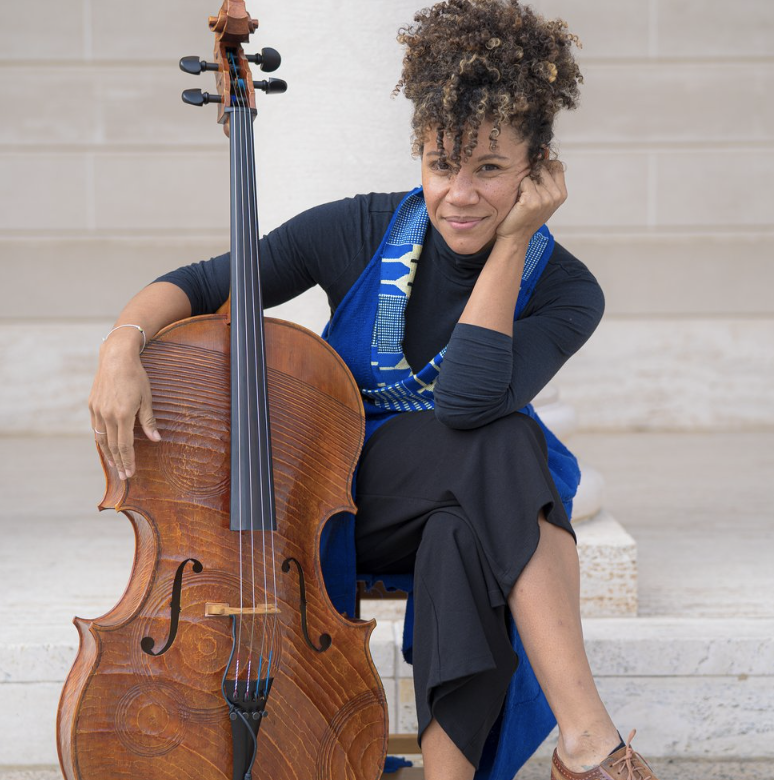 A woman sits, posing for a photo, holding her cello vertically. 
