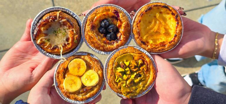 Hands holding several different varieties of egg tarts, topped variously with banana slices, pistachios, blueberries and more.