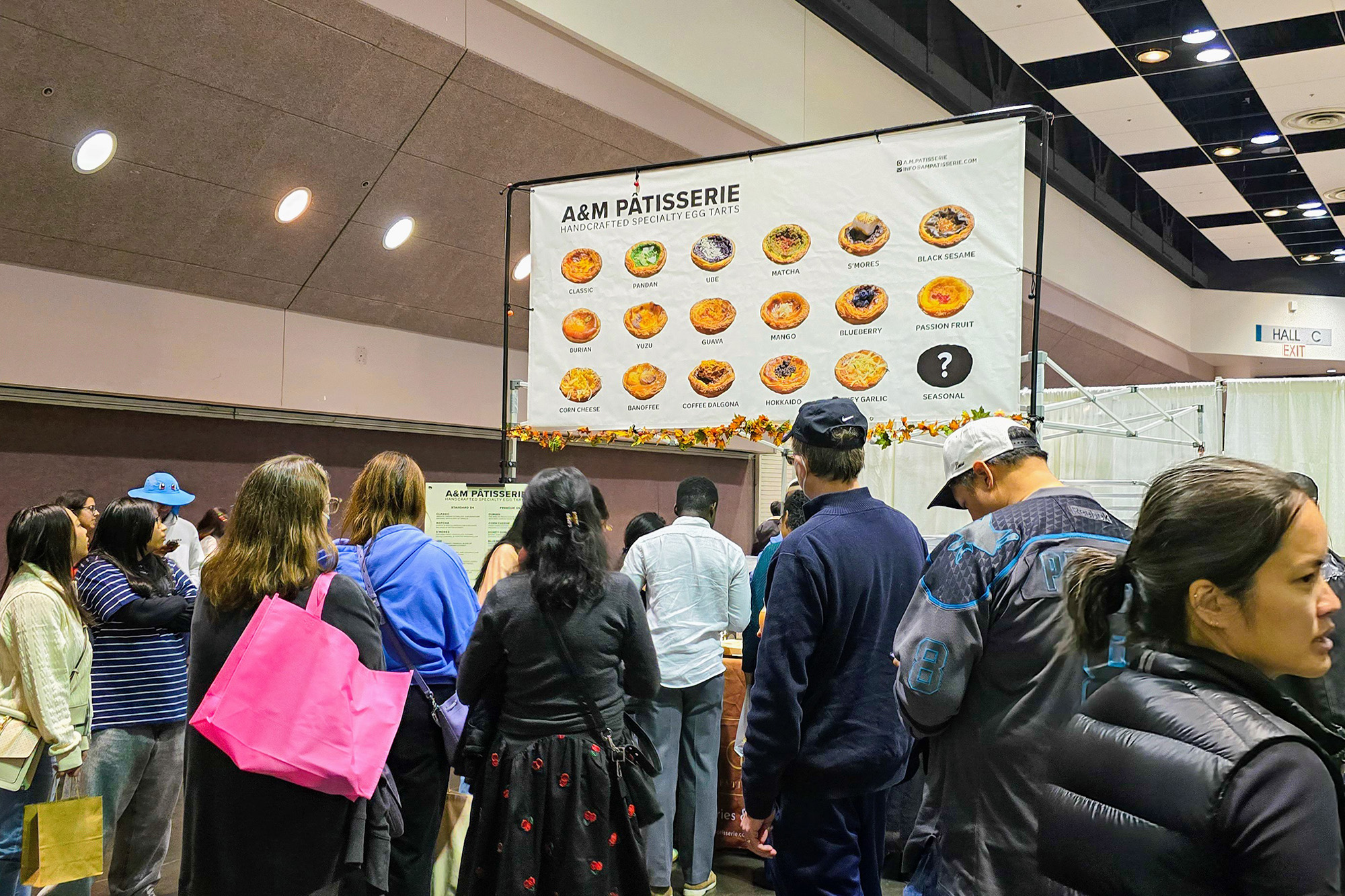 Long line of customers waiting to buy egg tarts. A large banner overhead shows the different flavors available.