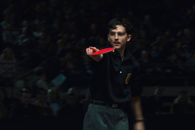 man points with hand holding table tennis paddle, blurry crowd behind
