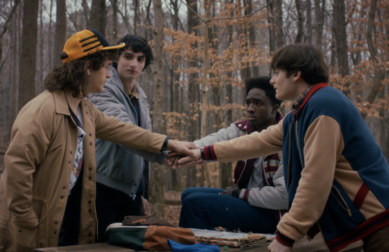 Four ’80s-era teens stand around a table in an autumnal forest. They all touch hands over the center of the table.