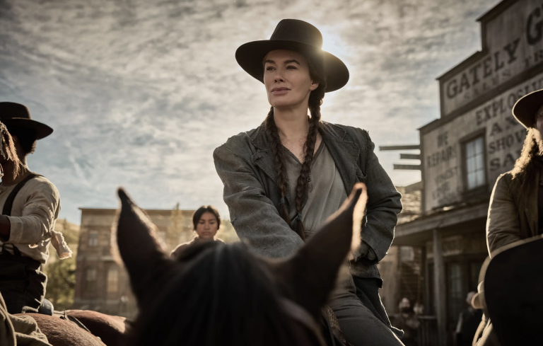 A white woman with long dark hair sits atop a horse, wearing a hat and practical Old West riding gear. She has a self-assured facial expression.