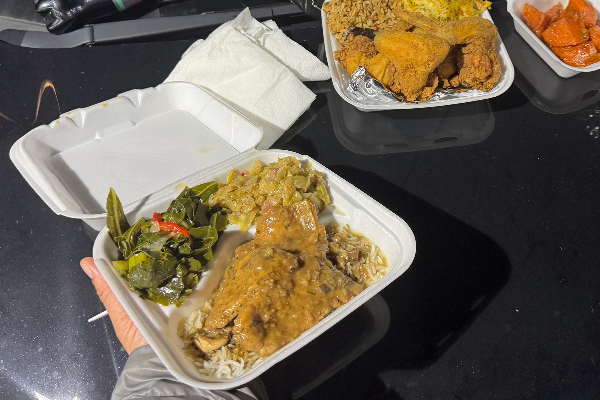 A smothered pork chop plate and other soul food dishes in styrofoam takeout containers, arranged on the hood of a car.