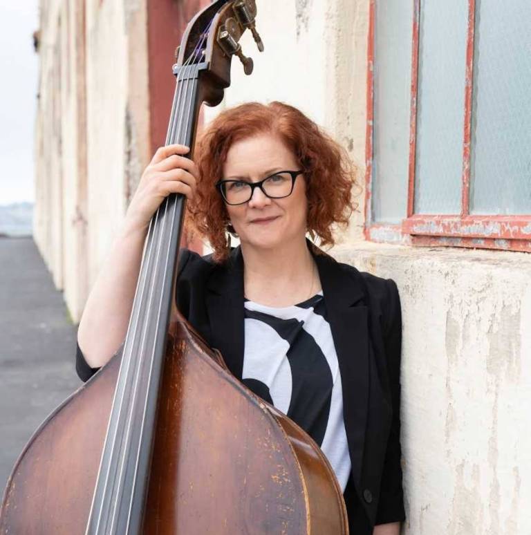A woman with glasses stands next to a building while posing for a photo, holding an upright bass.