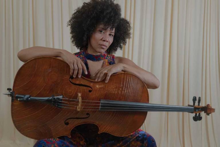 A woman sits and poses for the camera while holding a cello horizontally in her lap.