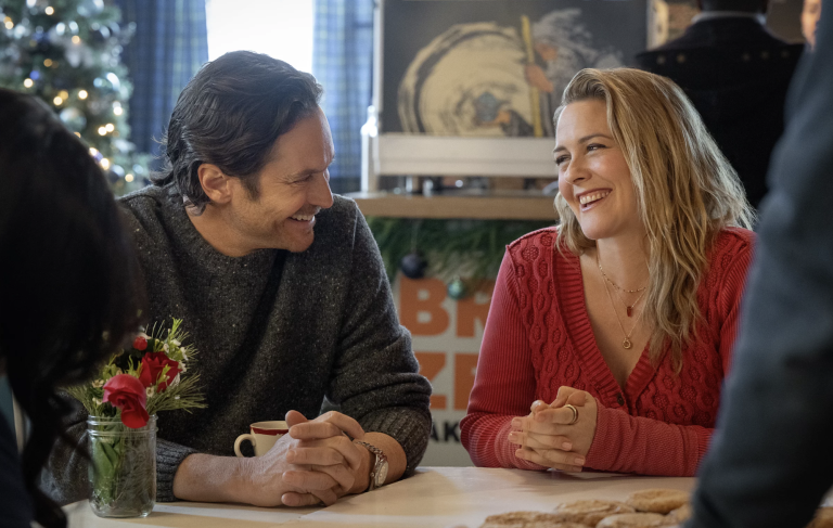 A middle aged white man and woman sit next to each other at a dining table. They are smiling warmly at one another and both wearing cosy sweaters.