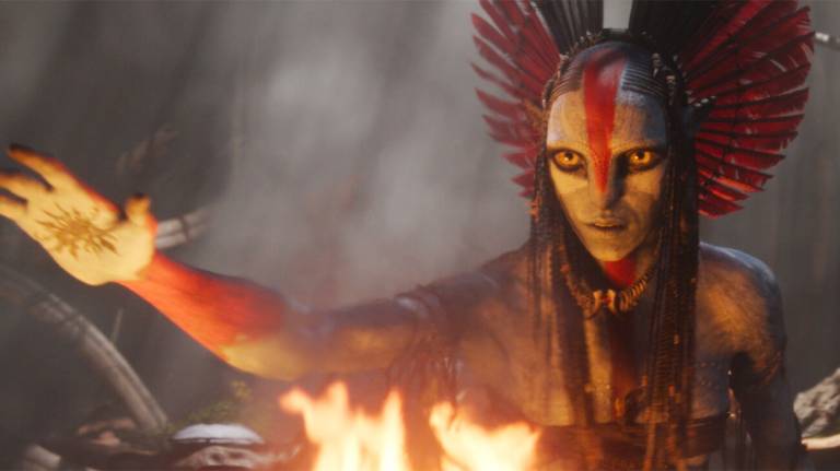 alien character with headdress raises hand over fire