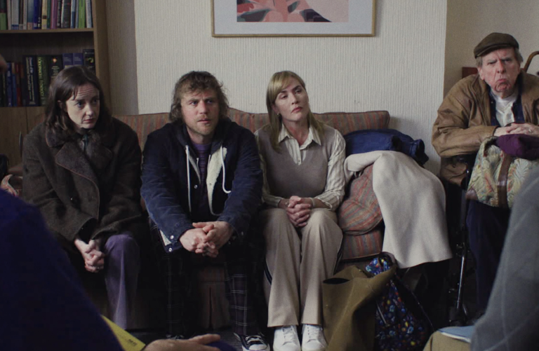 Two middle aged white women sit either side of a middle aged white man on a couch in an office. To their left is an older man sitting in a chair. Their facial expressions are a mixture of concerned and stoic.