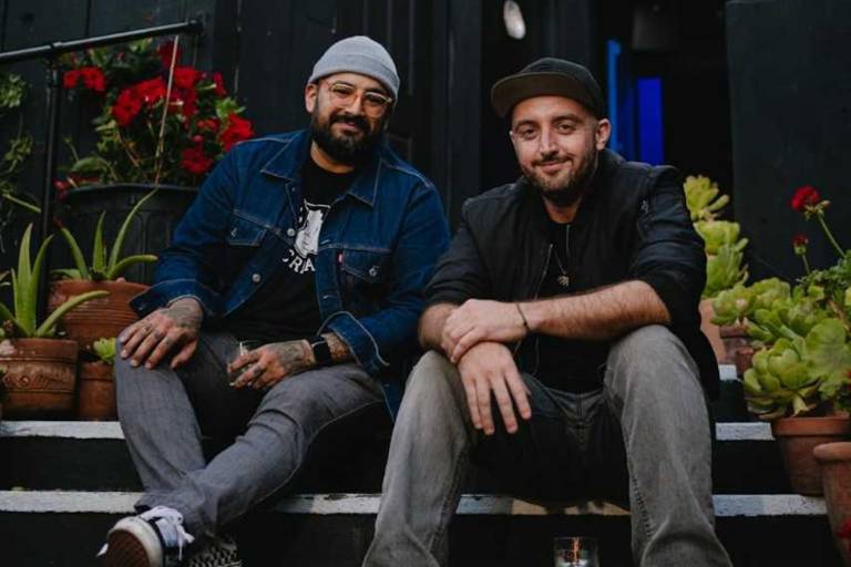 two men in beanies smile while sitting on steps, potted plants behind