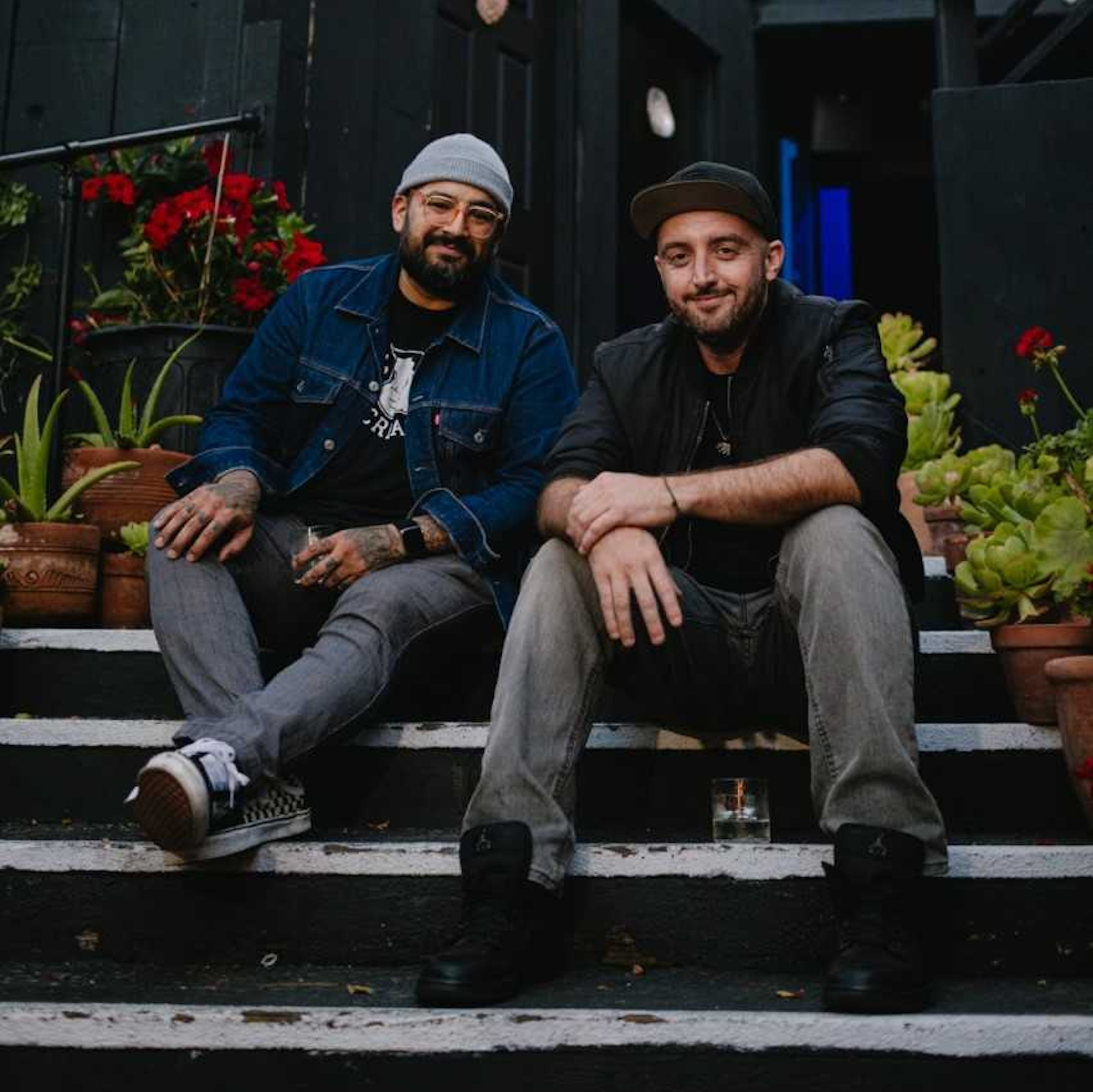 two men in beanies smile while sitting on steps, potted plants behind