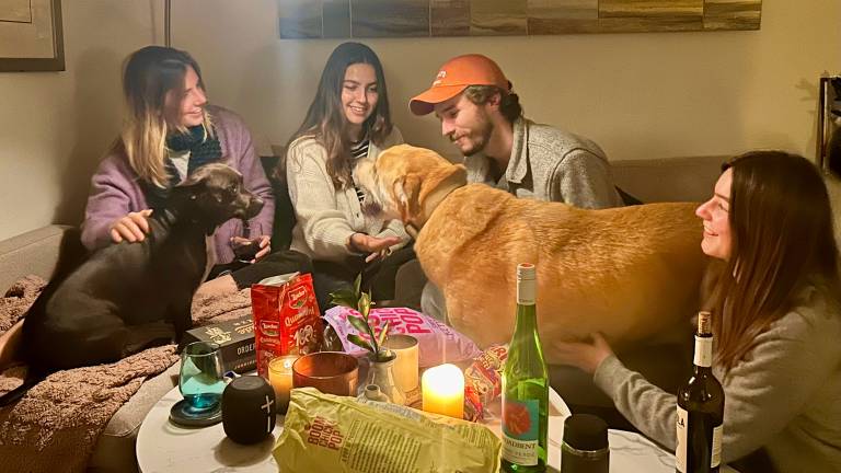 Four smiling people in their 20s and two dogs sit on the couch and floor to gather around a coffee table covered in wine, snacks, a plant and a candle.
