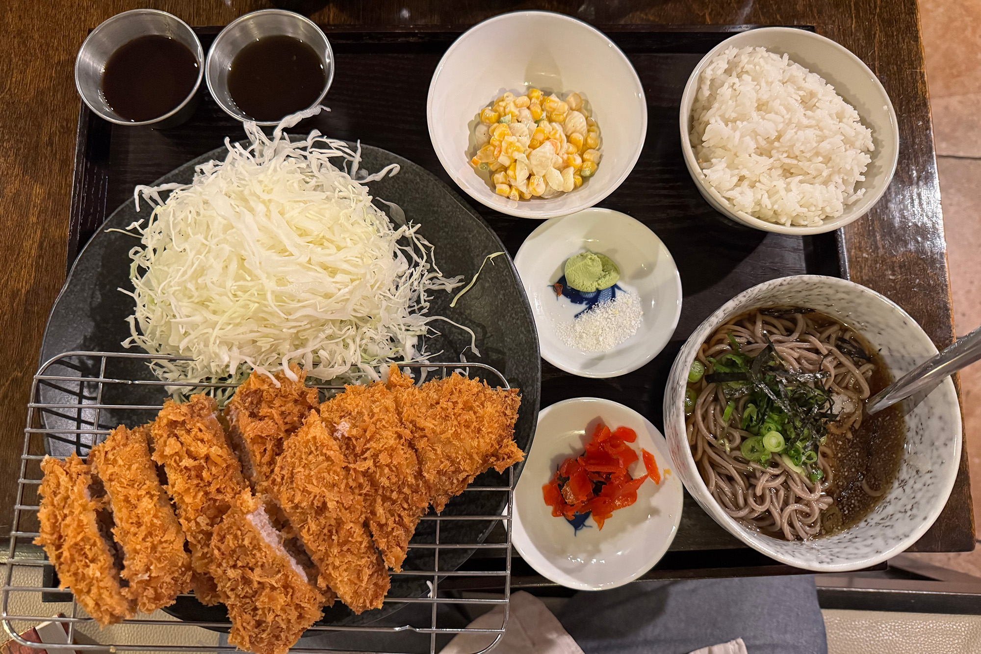 A Japanese-style fried pork cutlet served with raw cabbage, pickles, rice, and cold soba.