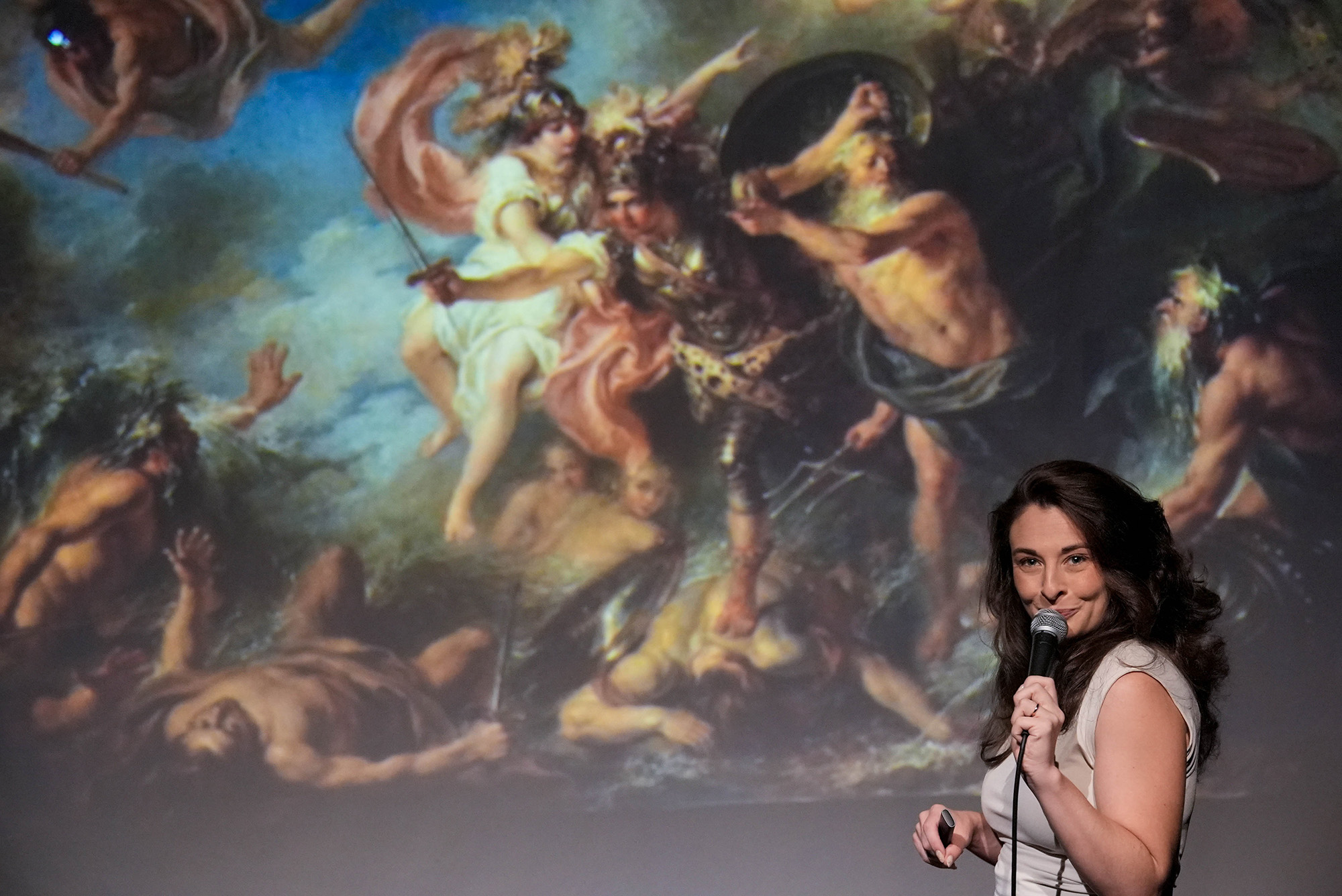 A woman holds a microphone in front of a projected image of a Renaissance painting.