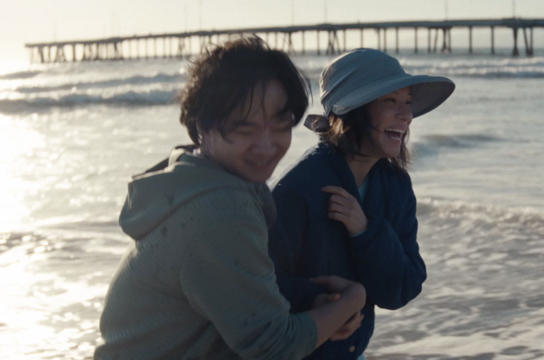 A young Asian man and a middle aged Asian woman cling to each other playfully as they take a windswept walk on the beach. The ocean and a pier are visible behind them.
