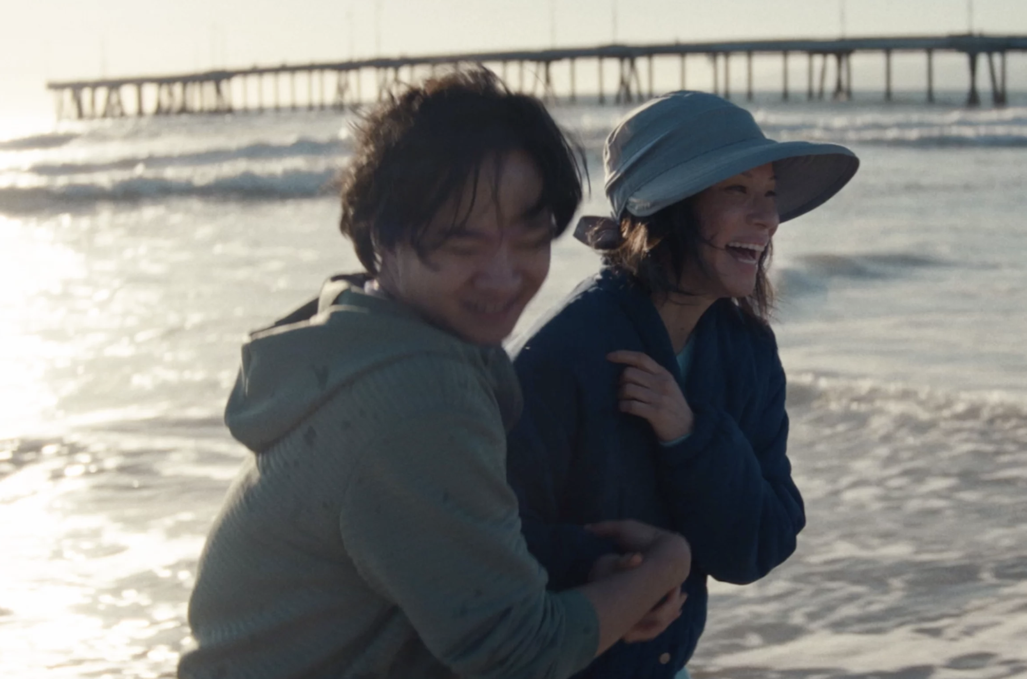 A young Asian man and a middle aged Asian woman cling to each other playfully as they take a windswept walk on the beach. The ocean and a pier are visible behind them.