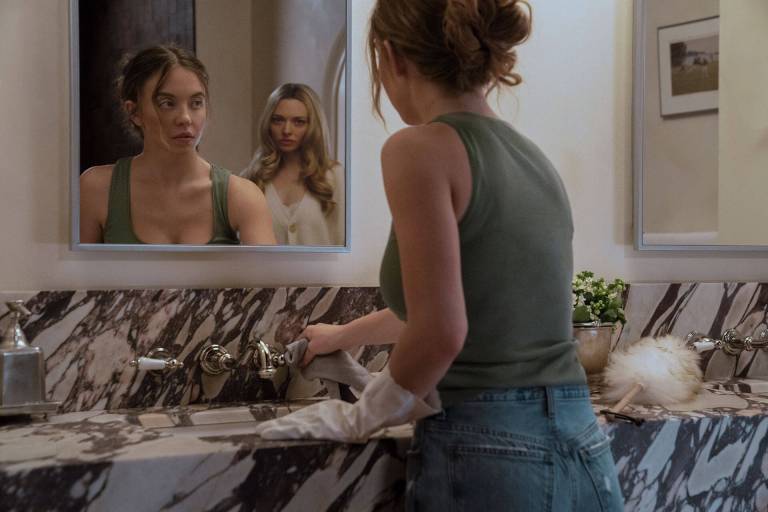 A woman cleaning a bathroom sink looks at another woman in the mirror reflection.