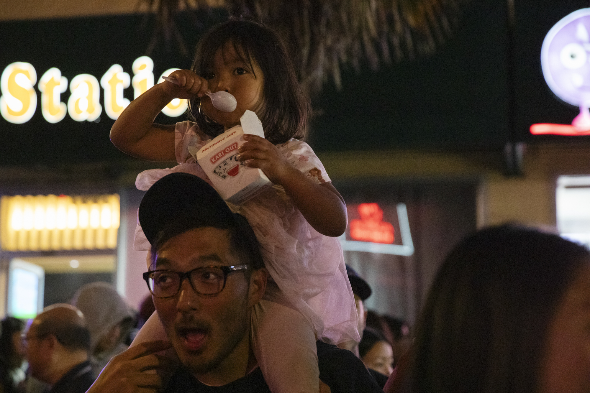 A father carries his young daughter on his shoulders as she eats food from a Chinese takeout carton.